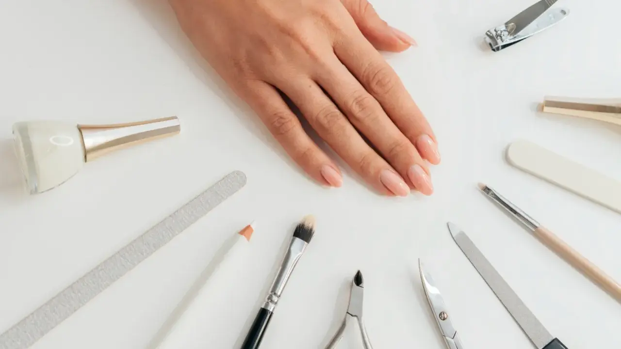 Dłoń z naturalnymi paznokciami otoczona akcesoriami do manicure. Wśród nich pilnik do paznokci naturalnych, cążki, nożyczki i pędzelki.