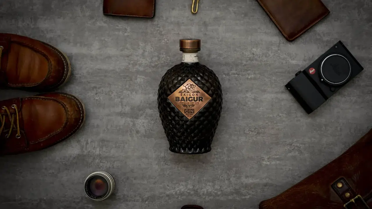 Saigon Baigur Gin mit 43% Alkoholgehalt, umgeben von Lederaccessoires, Kamera und Schuhen.