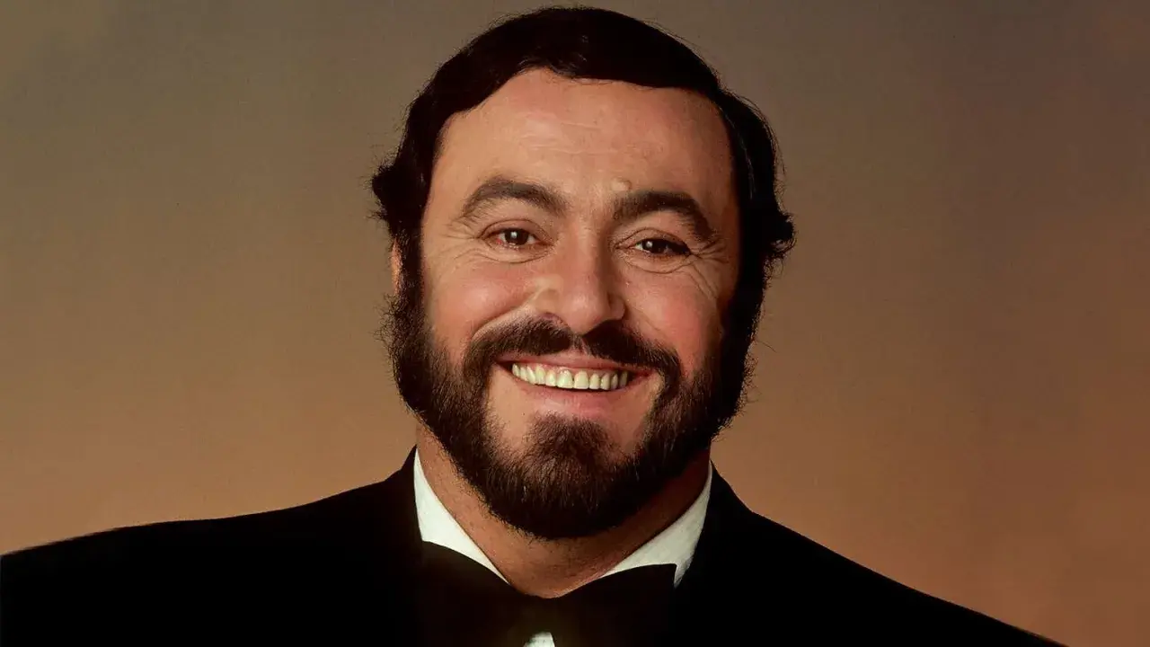 Uśmiechnięty Luciano Pavarotti w czarnym smokingu. Jego głos porusza serca, a jego uśmiech mówi "ti amo".