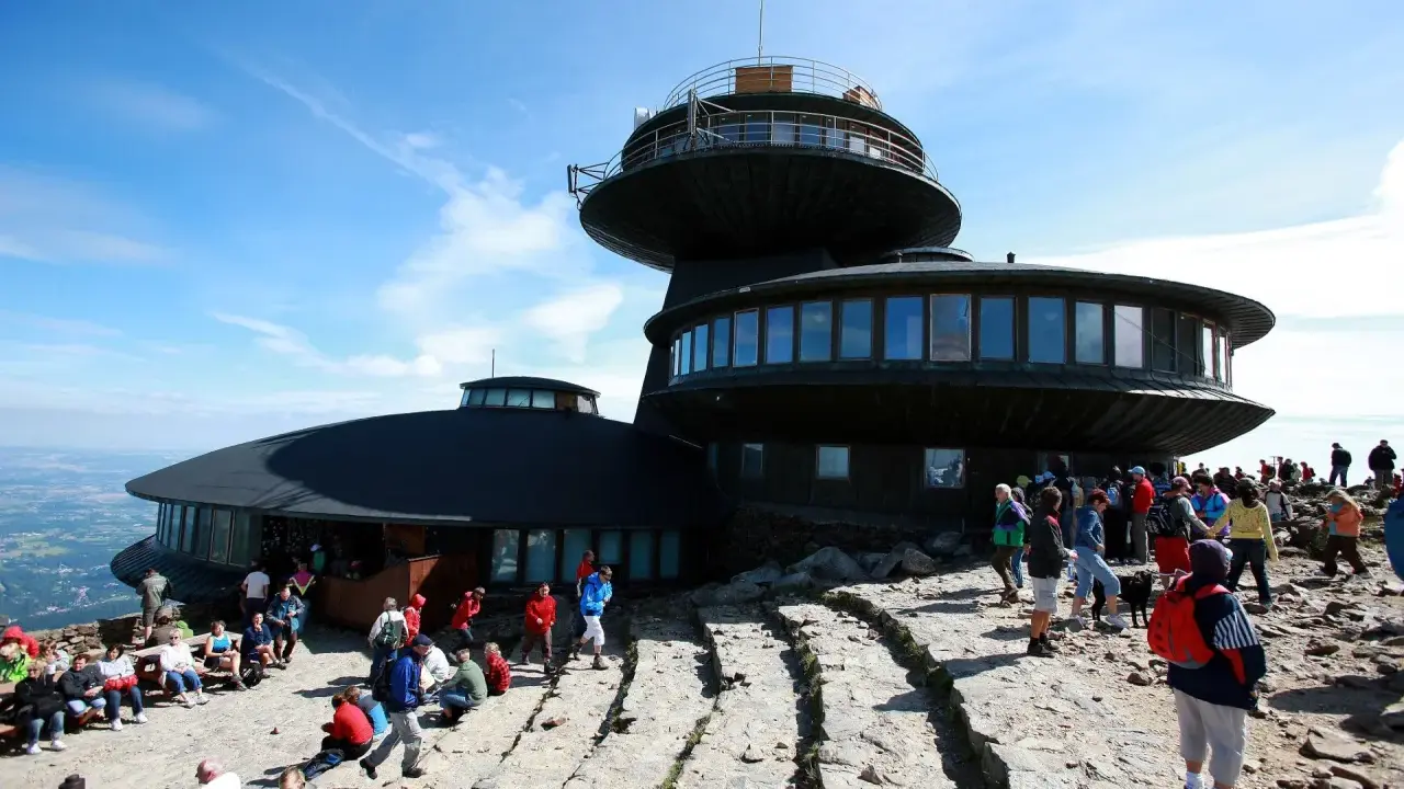 Ludzie odpoczywają na kamienistym szlaku na Śnieżkę, podziwiając widok na budynek obserwatorium.