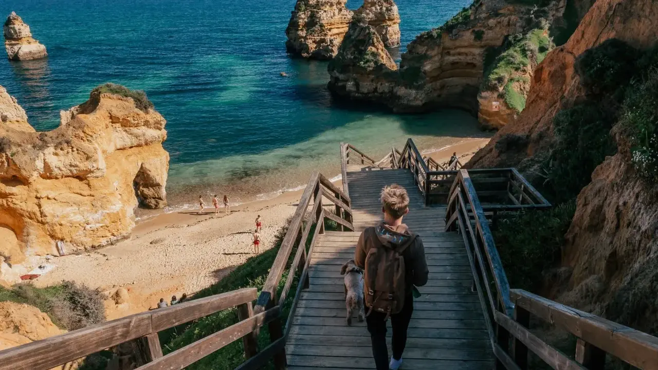 Persona desciende escaleras de madera hacia una hermosa playa de lagos con formaciones rocosas y aguas turquesas.