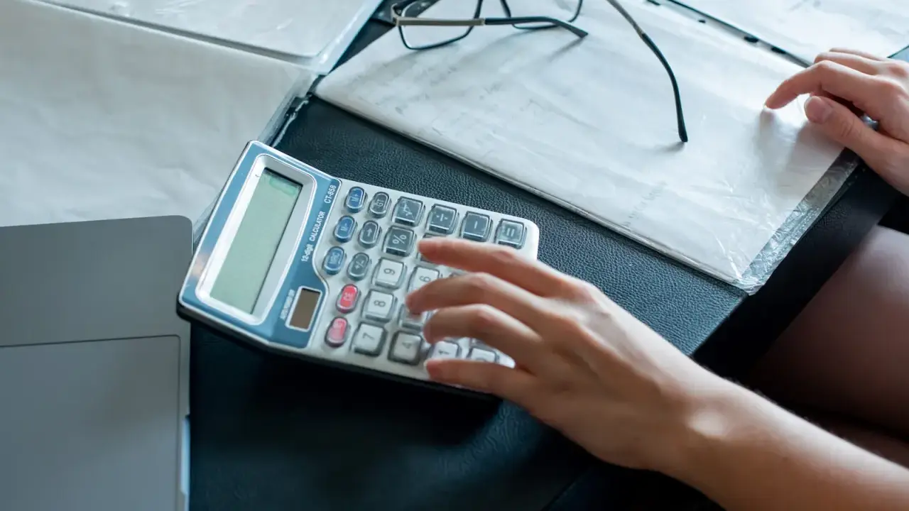 Ręce liczą na kalkulatorze dokumenty, analizując umowę zlecenie a alimenty. Obok leżą okulary i notatki.