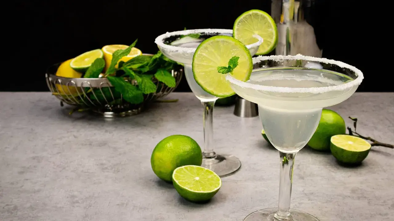 Zwei Margaritas mit Salzrand, garniert mit Limettenscheiben und Minze. Limetten und Minze in einer Schale im Hintergrund. Perfekt für ein Margarita Rezept.