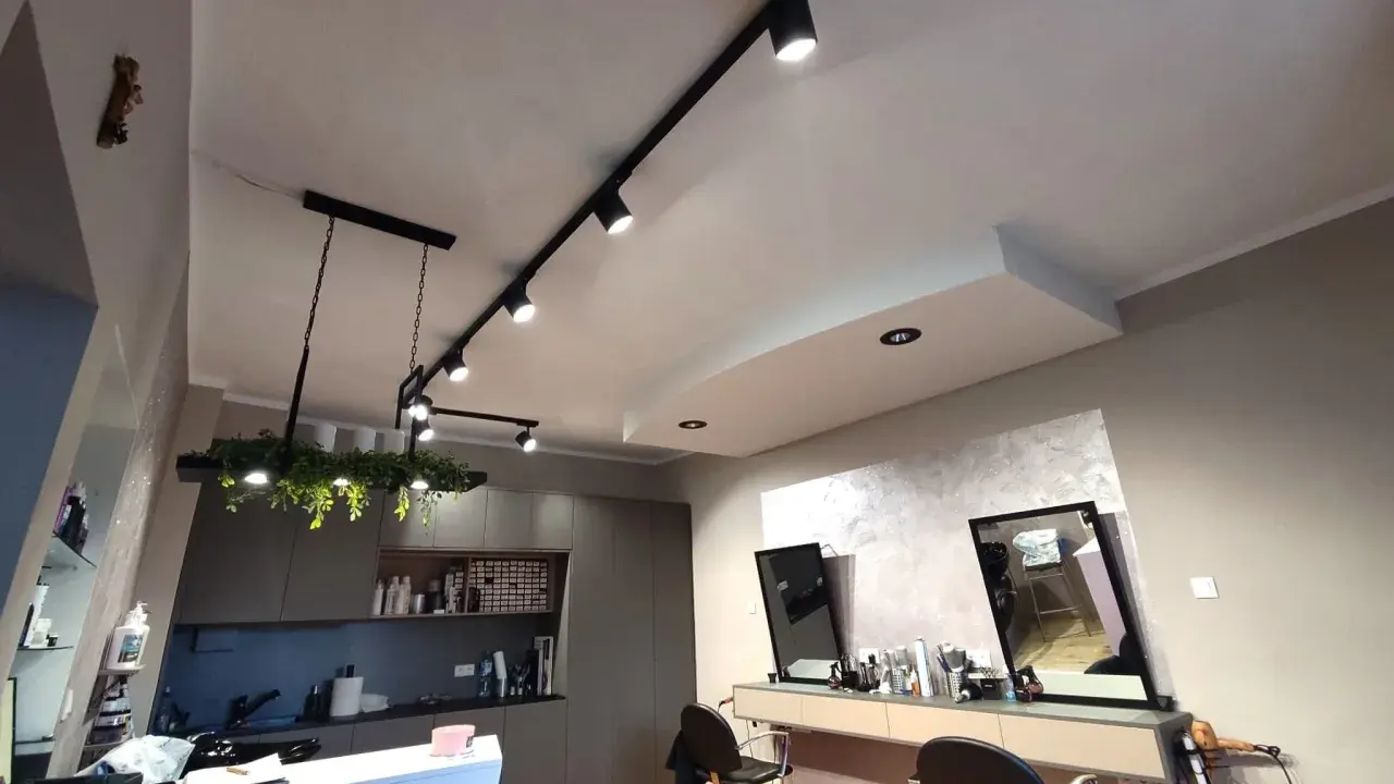 Nowoczesne oświetlenie w salonie fryzjerskim tworzy przytulną atmosferę. Reflektory na szynach i wisząca lampa z zielenią podkreślają styl wnętrza.