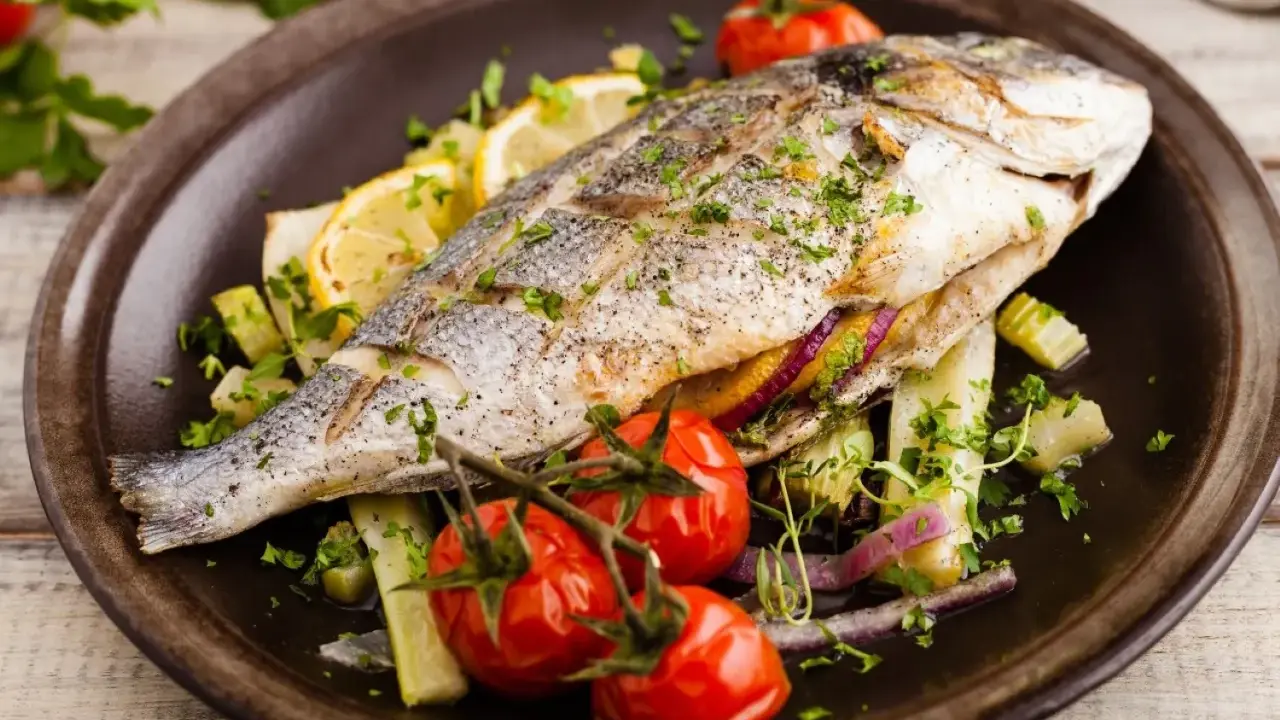 Poisson entier, probablement une truite au barbecue, servi avec des tomates cerises, des quartiers de citron et des herbes fraîches sur une assiette marron.
