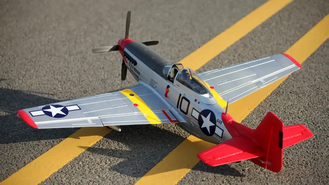 Ein detailgetreues Modell eines Mustang-Flugzeugs mit roten und silbernen Akzenten steht auf asphaltiertem Untergrund mit gelben Markierungen.