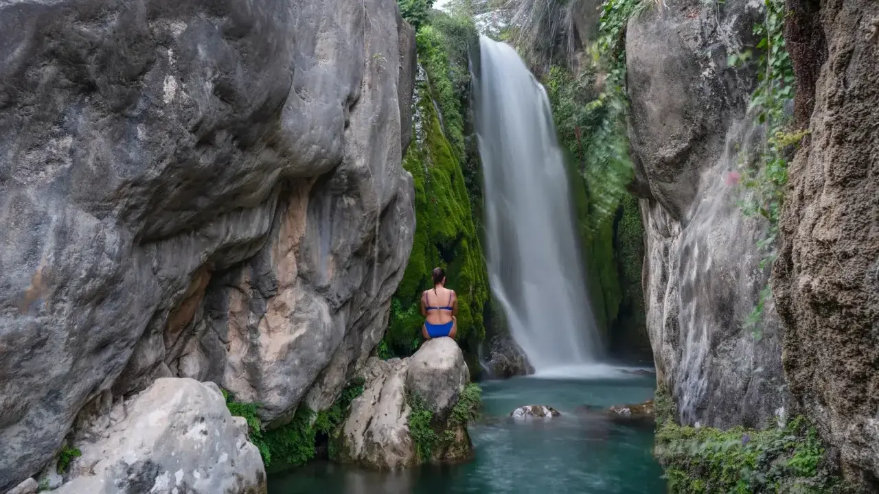 Una mujer sentada en una roca contempla la cascada de las fuentes del Algar, rodeada de vegetación y rocas.