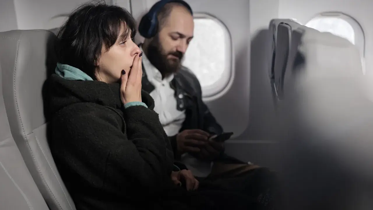 Kobieta z dłońmi na ustach, wyraźnie odczuwająca strach przed lataniem. Obok siedzi mężczyzna w słuchawkach.