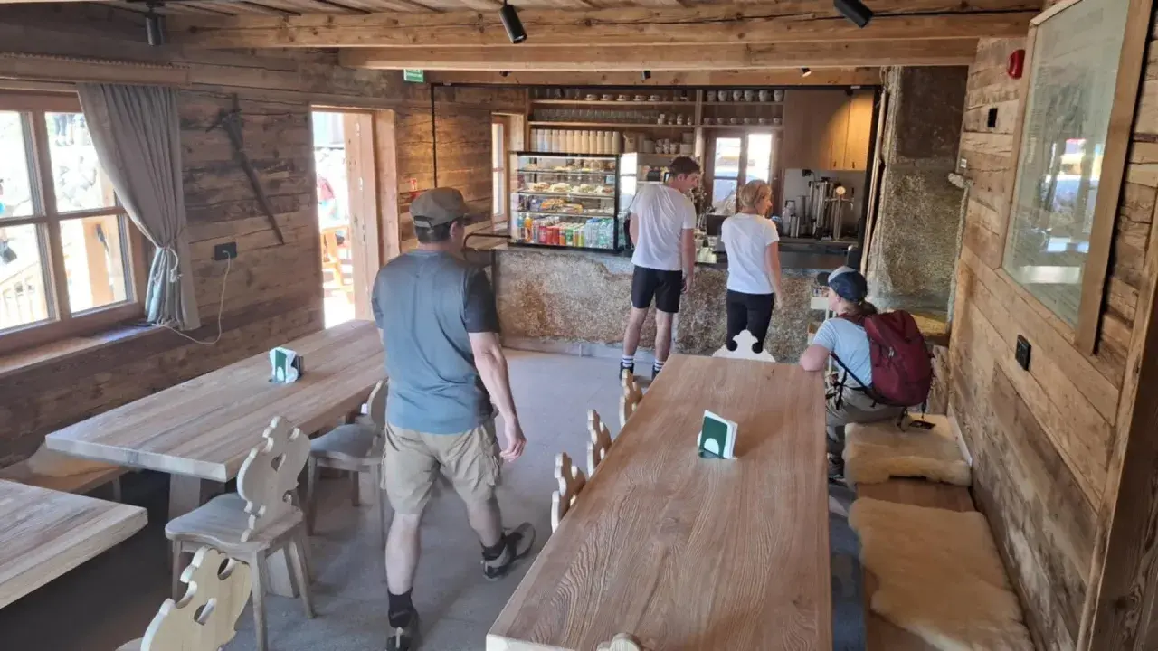 Wnętrze schroniska na Hali Kondratowej. Drewniane stoły i ławy, bar z ekspozycją ciast i napojów. Kilka osób odpoczywa po wędrówce.