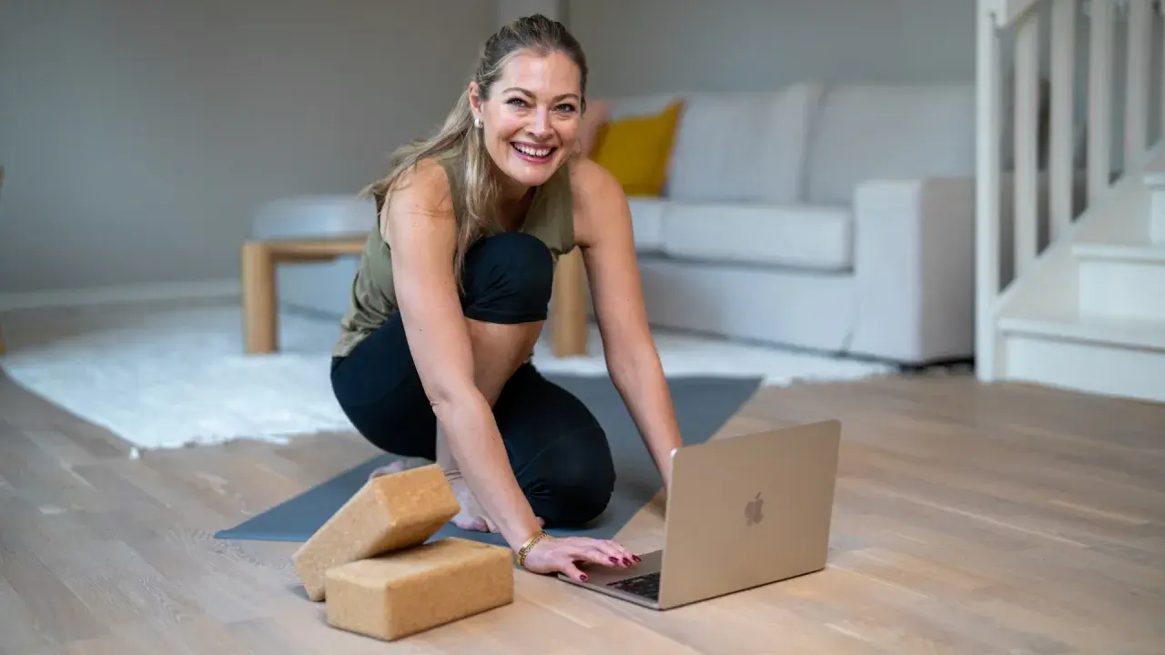 Glad kvinna förbereder sig för en lugn stund med **restorative yoga** hemma, med en laptop och yogablock redo.