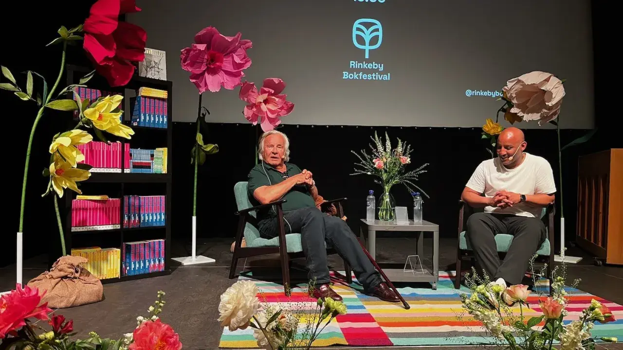 Jan Guillou delar fakta på Rinkeby Bokfestival. Scenen är dekorerad med stora blommor och bokhyllor.