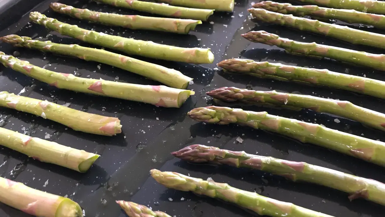 Färska sparrisar ligger på en ugnsplåt, redo att rostas i ugnen. Saltkorn glittrar på de gröna stjälkarna.