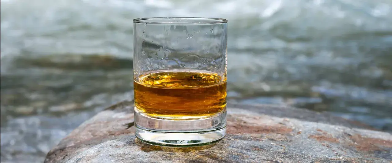 Ein Glas goldener Whisky steht auf einem Stein am Flussufer. Die ideale trinktemperatur für diesen edlen Tropfen.