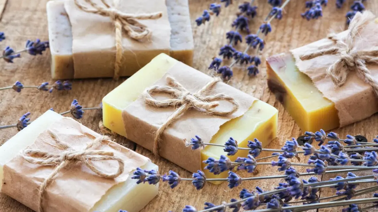 Quattro saponette fatte in casa, avvolte in carta e legate con spago, giacciono su un tavolo di legno rustico, circondate da rametti di lavanda profumata.