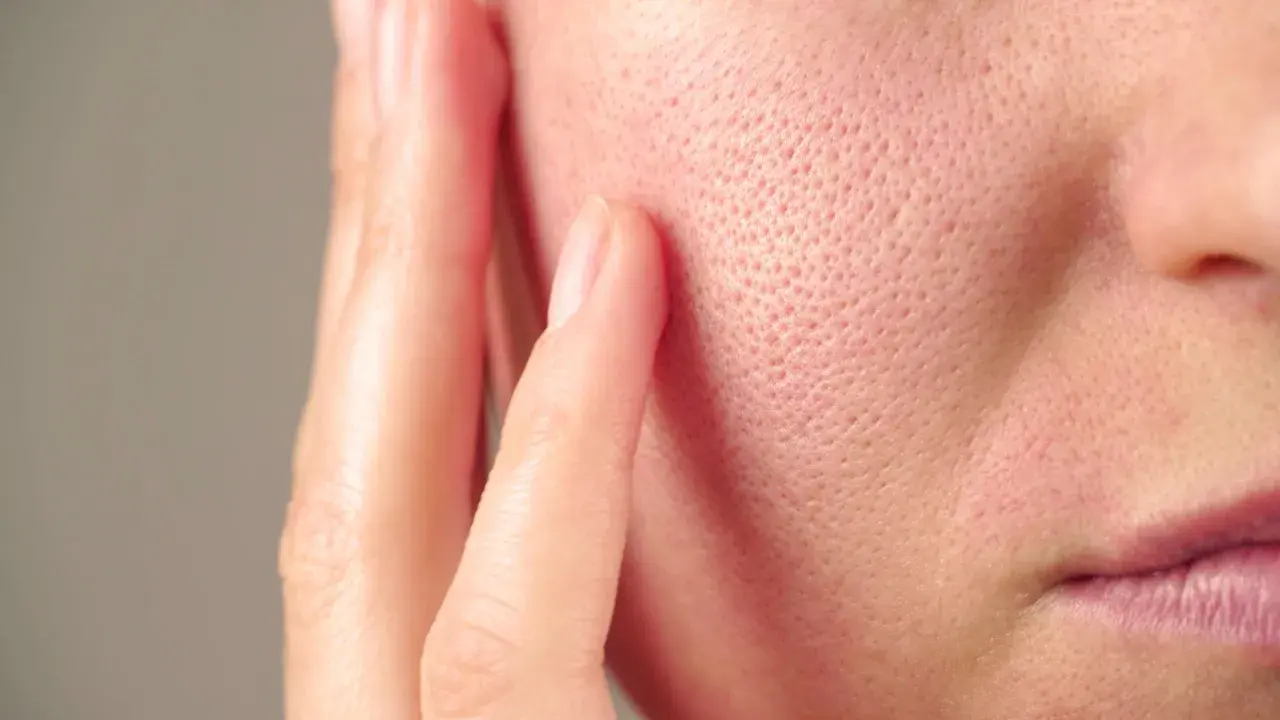 Gros plan sur la joue d'une personne, montrant les pores de la peau. Des doigts effleurent délicatement la surface.