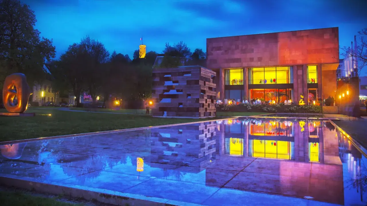 Abendstimmung am Museum mit reflektierendem Wasserbecken. Ein Turm leuchtet in der Ferne. Entdecke Bielefeld Geheimtipps!