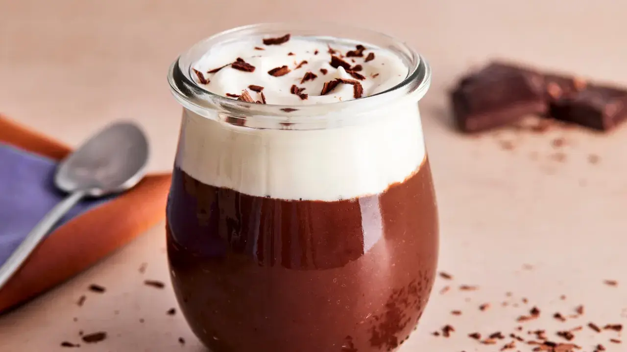 Ein Glas mit Omas Schokoladenpudding mit Sahne und Schokostreuseln. Daneben liegen ein Löffel und ein Stück Schokolade.