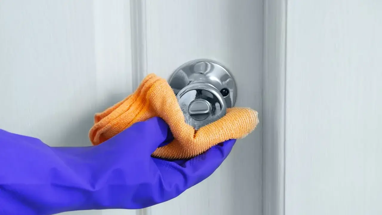 Ręka w fioletowej rękawiczce czyści klamkę pomarańczową ściereczką.