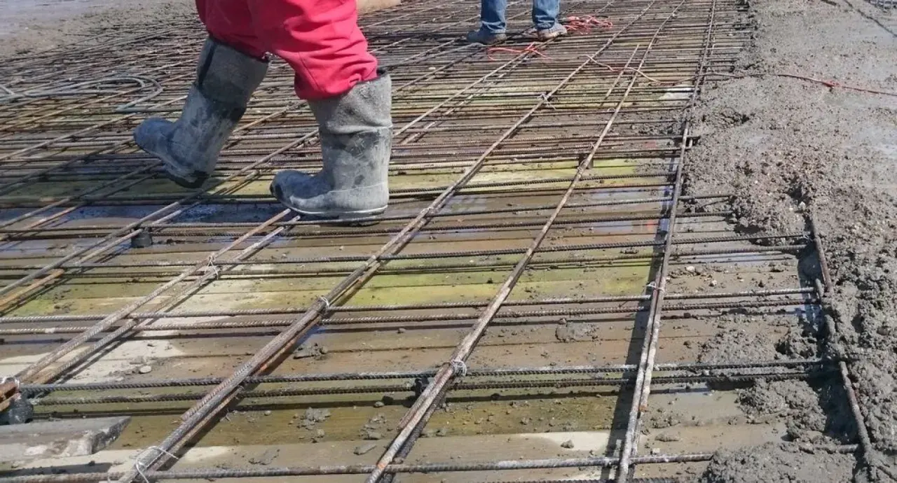 Pracownicy w gumowcach stąpają po siatce zbrojeniowej, przygotowując zbrojenie stropu do zalania betonem.