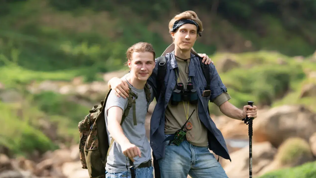 Dwóch młodych mężczyzn, gotowych na trekking, z plecakami i kijkami, w otoczeniu zieleni i skał.