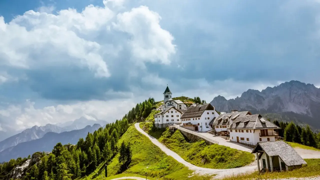 Un borgo incantevole in montagna, perfetto per le tue vacanze in Friuli. Case bianche, una chiesa e sentieri verdi sotto un cielo nuvoloso.