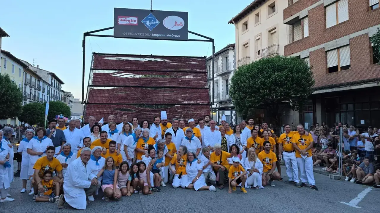 Grupo posa en la fiesta longaniza Graus, con un cartel de "Los Tres Sabores de la Longaniza de Graus" y una estructura de madera con embutidos.