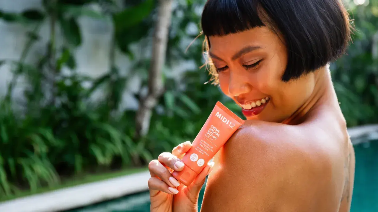 Une femme souriante applique une crème solaire SPF 50+ sur son épaule. Le tube indique "Très haute protection", le SPF 50 signification est une protection optimale.