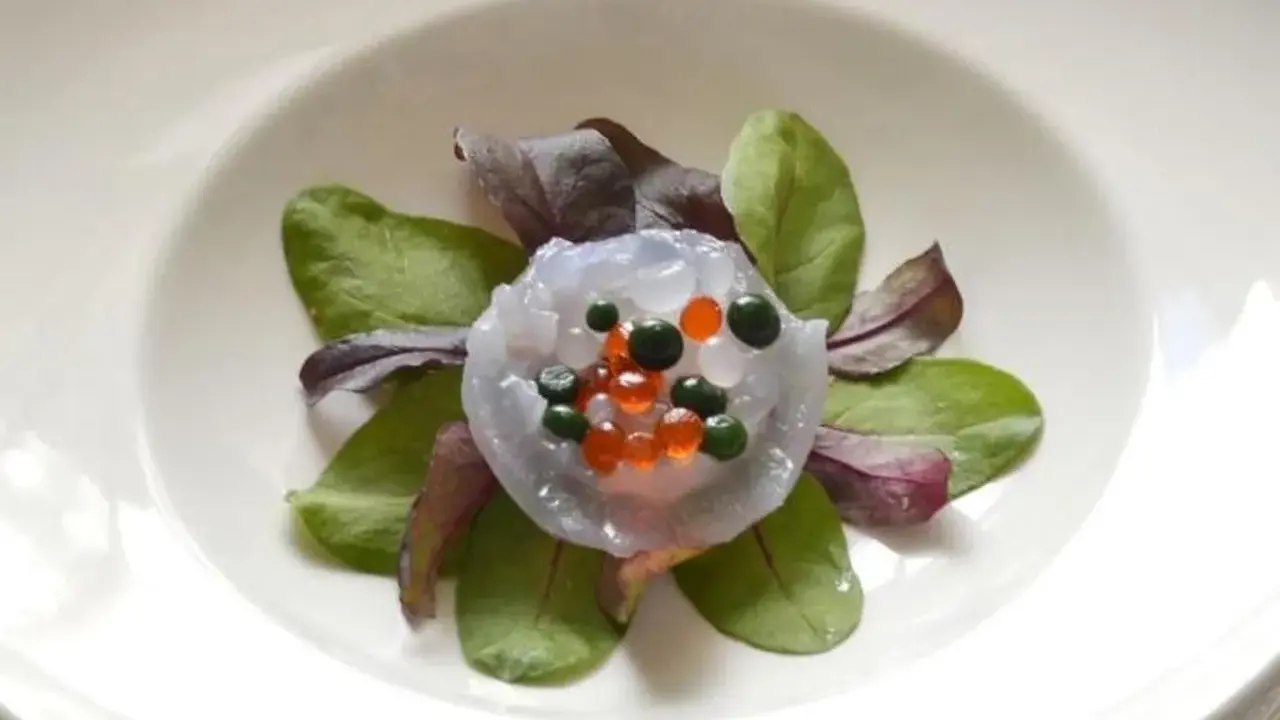 Piatto elegante con meduse commestibili trasparenti, guarnite con sfere arancioni e verdi, su un letto di foglie verdi e rosse.