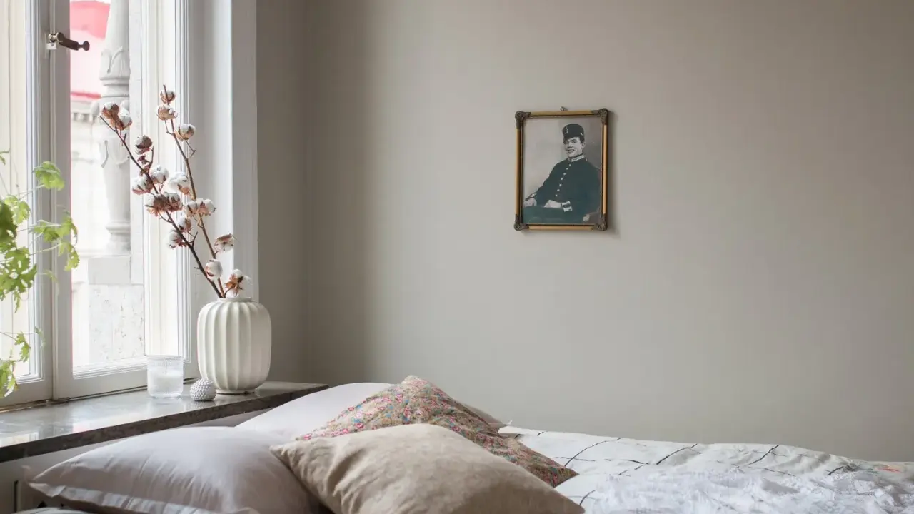 En säng med flera kuddar i olika nyanser av beige och mönster. En gammal porträttbild hänger på väggen.