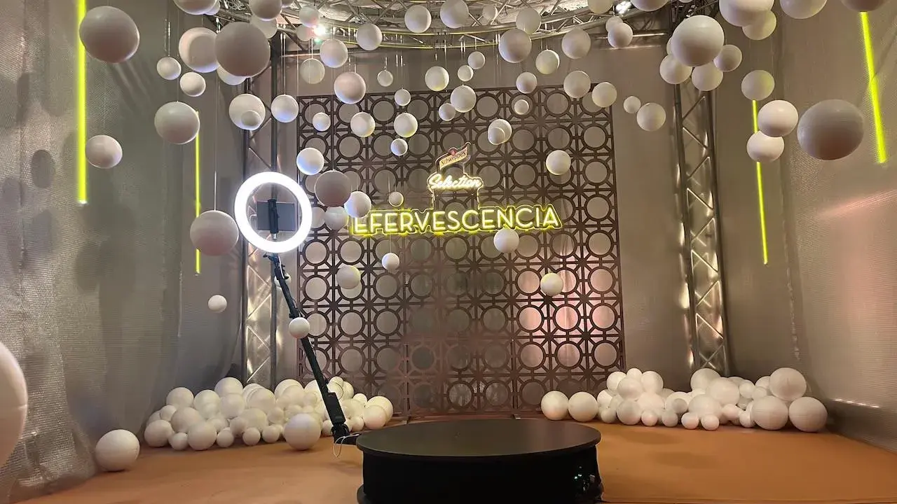 Plataforma giratoria para fotos con luces de neón "EFERVESCENCIA" y esferas blancas colgantes, creando un efecto de bullet time.