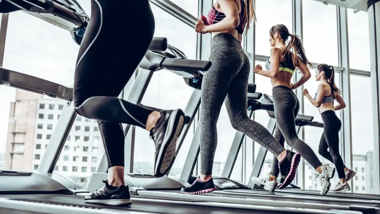 Kobiety ćwiczą cardio na bieżni, biegnąc w siłowni z widokiem na miasto.
