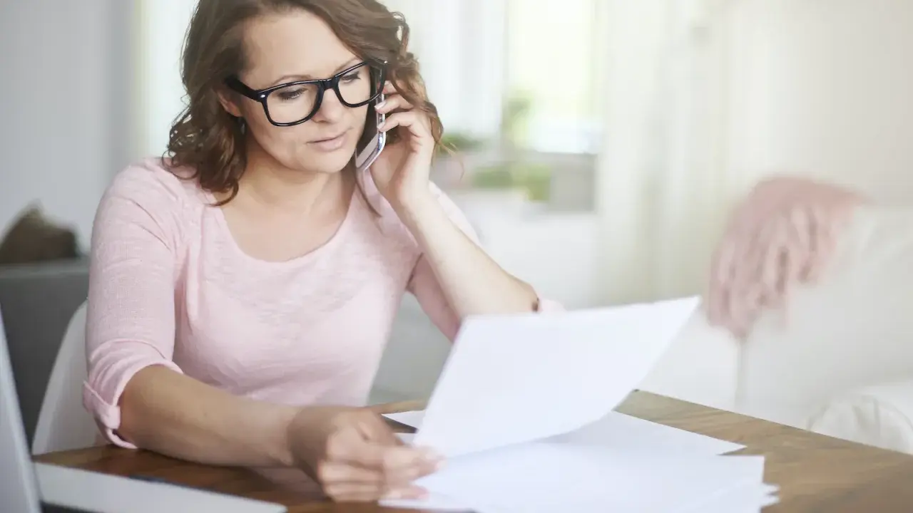 Kobieta w okularach rozmawia przez telefon, przeglądając dokumenty. Martwi się, że czeka ją brak świadectwa pracy.