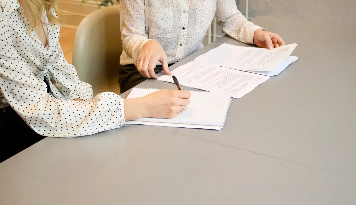 Dwie kobiety przy stole omawiają dokumenty. Jedna pisze, druga przegląda papiery. Może to umowa o zakazie konkurencji.