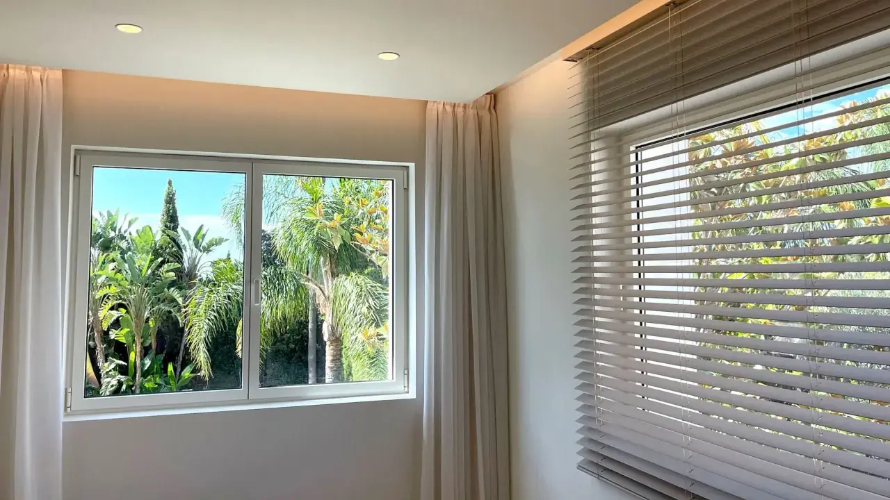 Dos tipos de apertura de ventanas: una corredera con cortinas y otra con persianas venecianas, ambas con vistas a un exuberante jardín tropical.