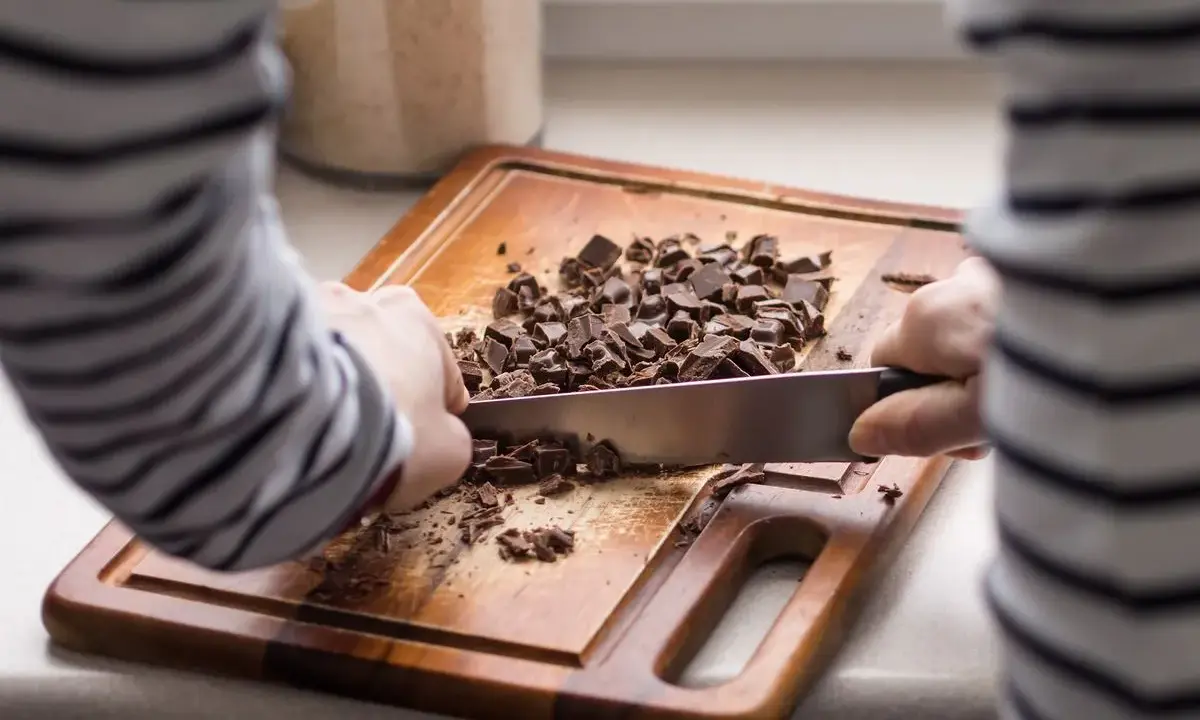 Une personne utilise un couteau pour comment casser une tablette de chocolat en pépites sur une planche à découper en bois.
