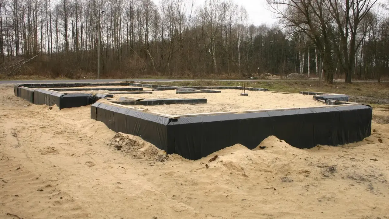 Fundament domu w piasku, otoczony czarną folią. W tle las. To początek budowy, gdzie dysperbent chroni fundament.