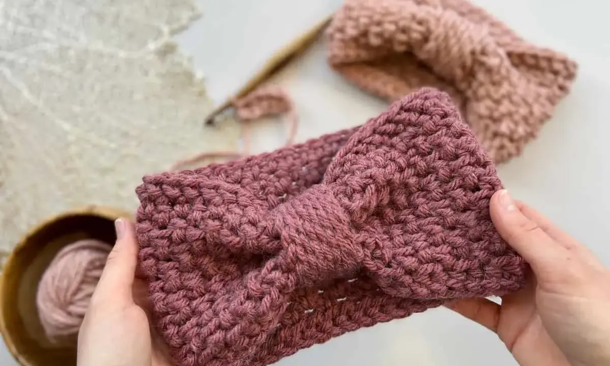 Manos sostienen una diadema tejida a crochet con un lazo. Detrás, otra diadema y ovillo de lana. ¡Descubre patrones gratis de diademas de ganchillo!