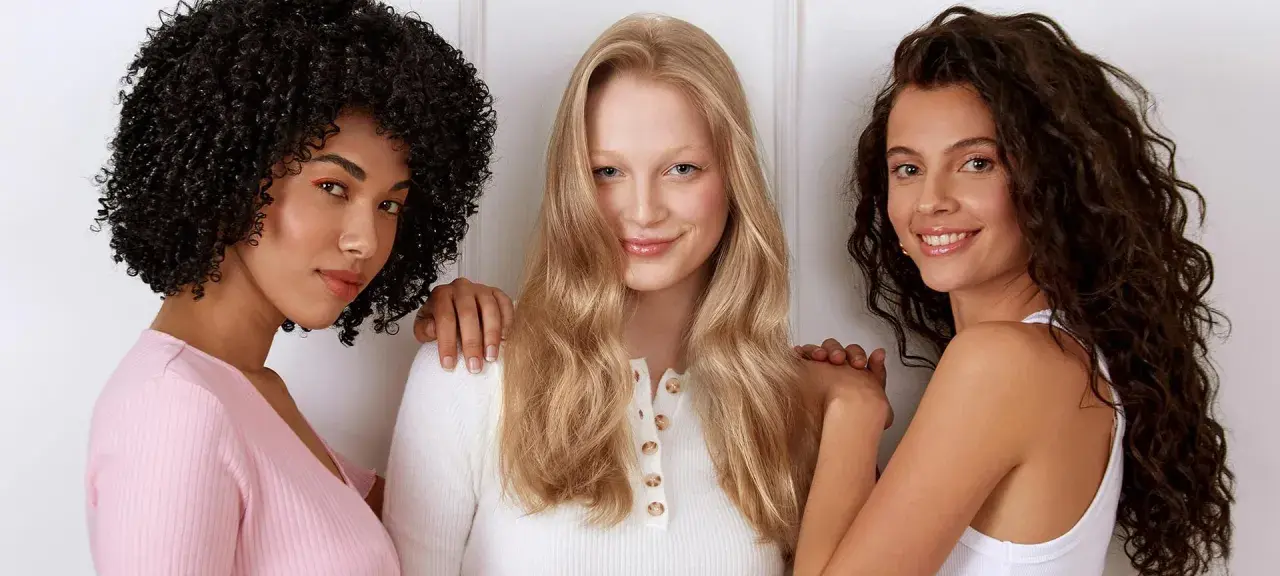 Drei Frauen stehen nebeneinander. Die Frau in der Mitte hat orange blonde haare und lächelt.