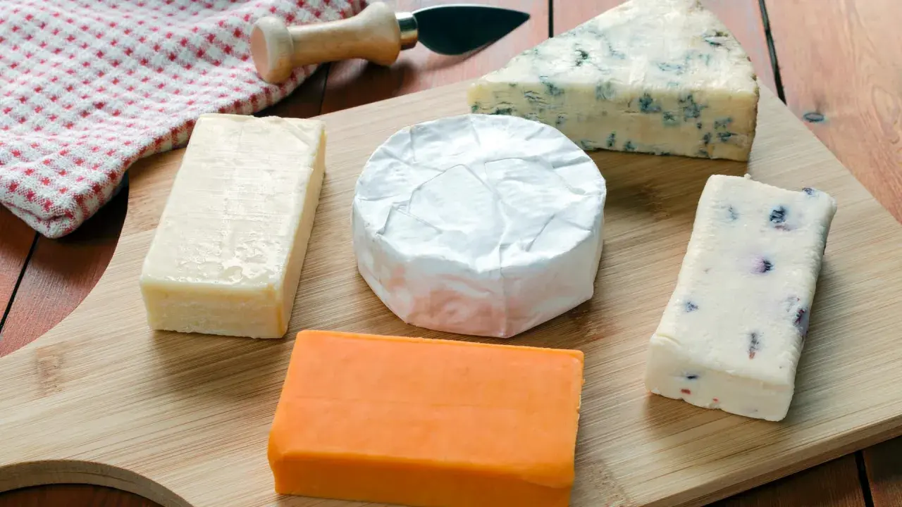 Verschiedene Käsesorten auf einem Holzbrett. Warum ist Cheddar orange? Die natürliche Farbe kommt von Beta-Carotin, das Kühe über Gras aufnehmen.