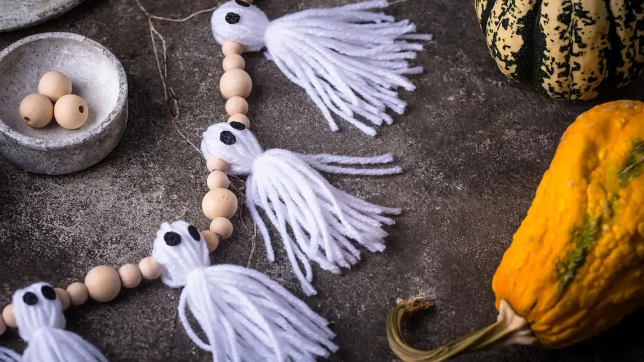Niedliche Geister-Girlande zum Halloween basteln, mit Holzperlen und weißen Fransen. Daneben liegen bunte Kürbisse.