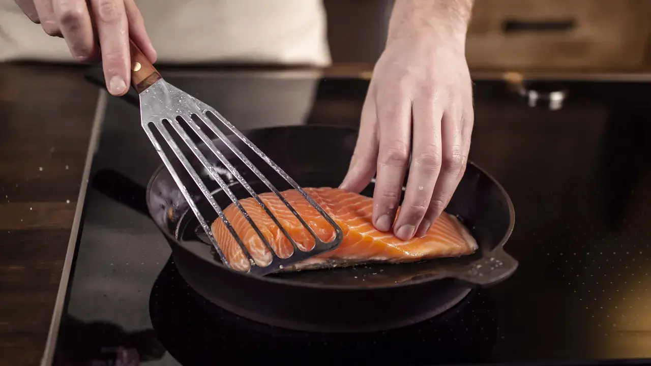 Ein Lachsfilet wird mit einem Pfannenwender in eine heiße Pfanne gelegt. Perfekt für leckere Fisch Rezepte!