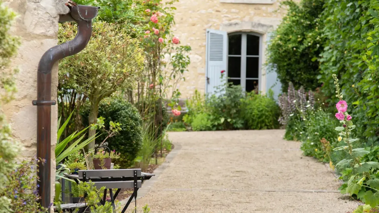 Un chemin de gravier serpente à travers un jardin anglais romantique, bordé de roses roses et de verdure luxuriante. Une maison en pierre avec des volets bleus se profile à l'arrière.