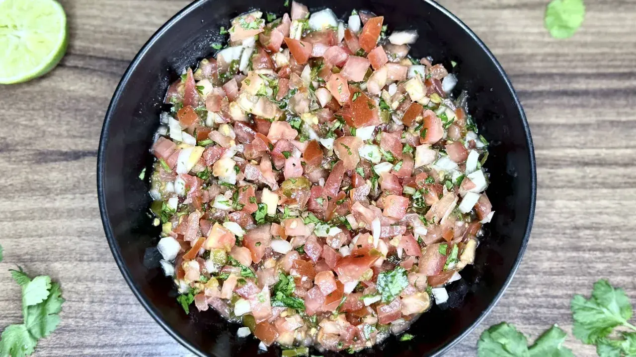 Świeże, kolorowe pico de gallo w czarnej misce, z limonkami i kolendrą na drewnianym tle.