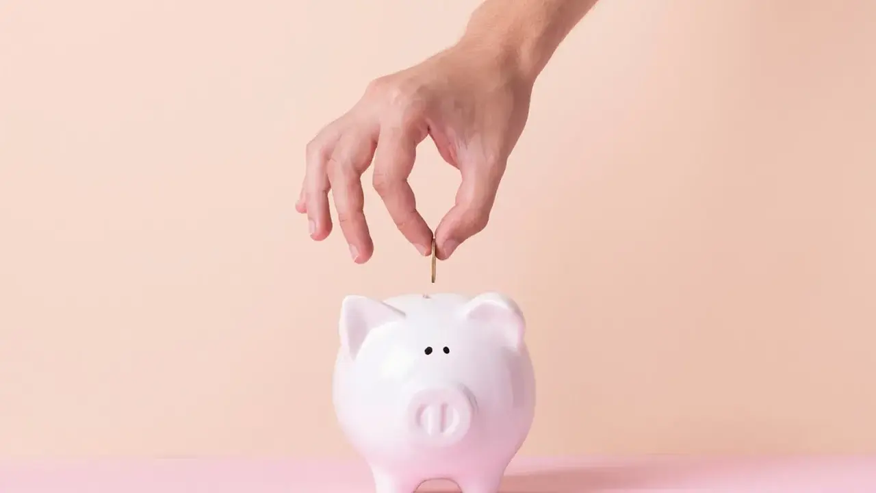 Mano inserta moneda en alcancía blanca, simbolizando cómo empezar una cuenta de ahorro.