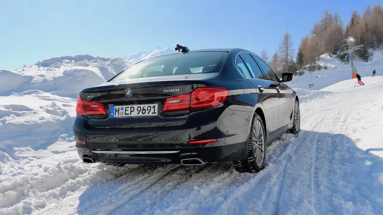 Schwarzer BMW 540i G30 fährt durch tiefen Schnee auf einer Skipiste, bergauf mit Skilift im Hintergrund.