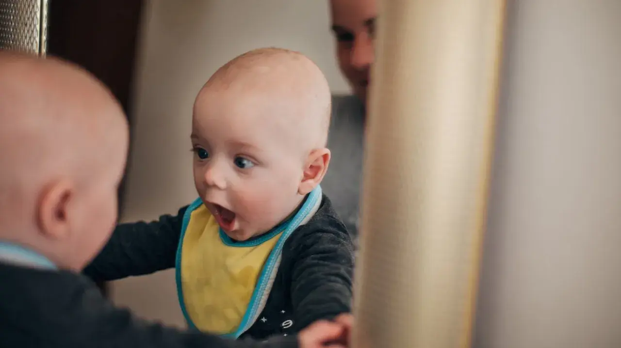 Ein Baby mit gelbem Lätzchen schaut überrascht in einen Spiegel. Vielleicht fragt es sich, ab wann Kinder "Ich" sagen.