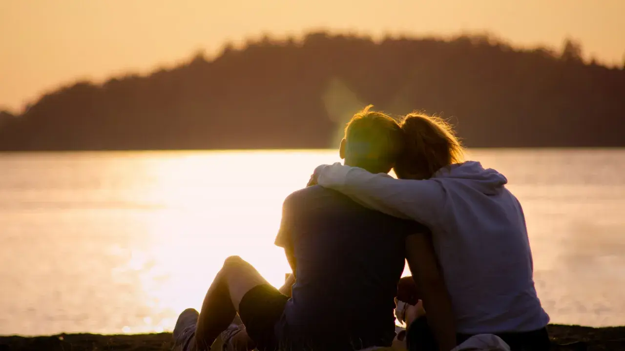 Ein Paar sitzt am Seeufer im Sonnenuntergang, die Arme umeinander geschlungen. Sie finden Trost und lernen, sich selbst zu verzeihen.