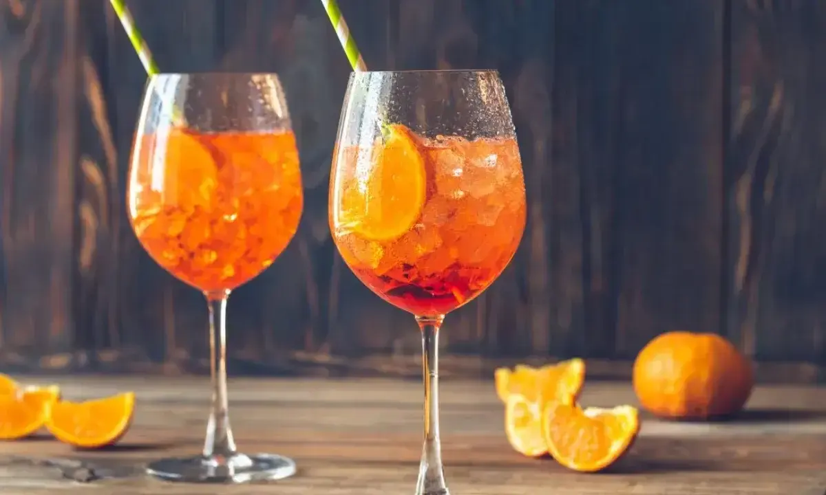 Dwa orzeźwiające Aperol Spritz z plasterkami pomarańczy i słomkami, na drewnianym stole.