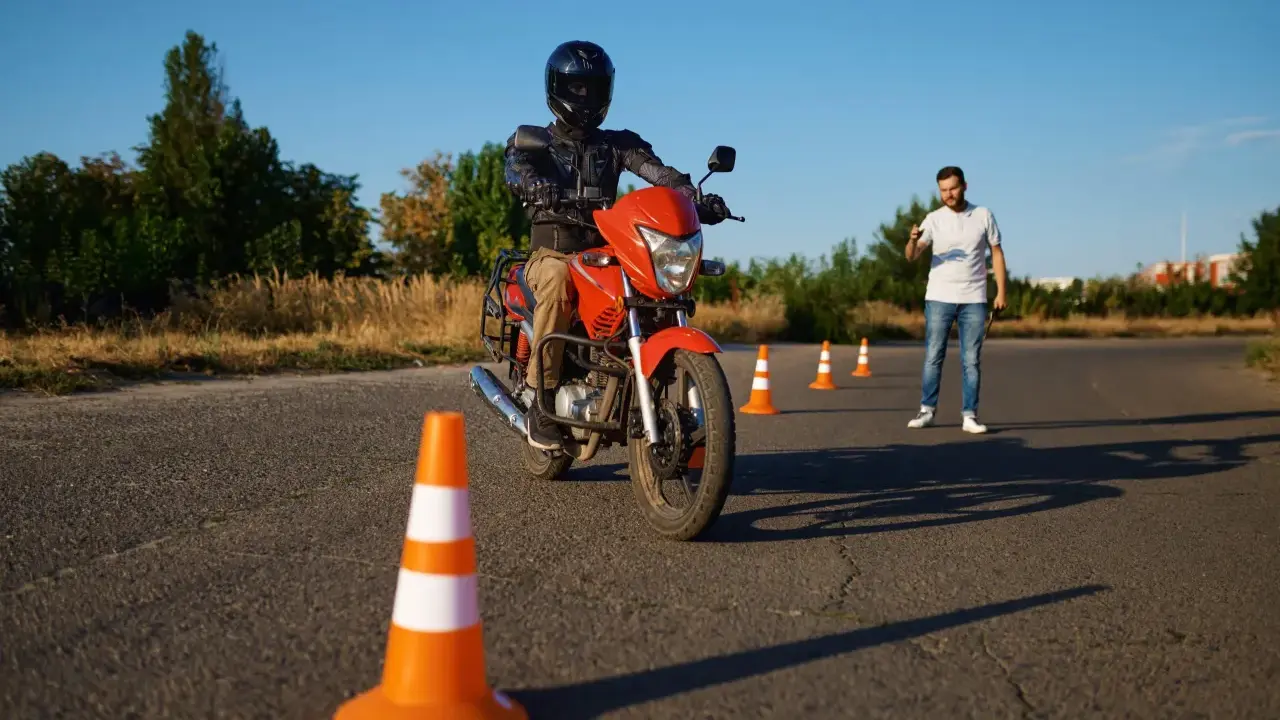 Persona con casco y equipo de protección conduce una moto naranja en un circuito de prácticas. Un instructor observa.