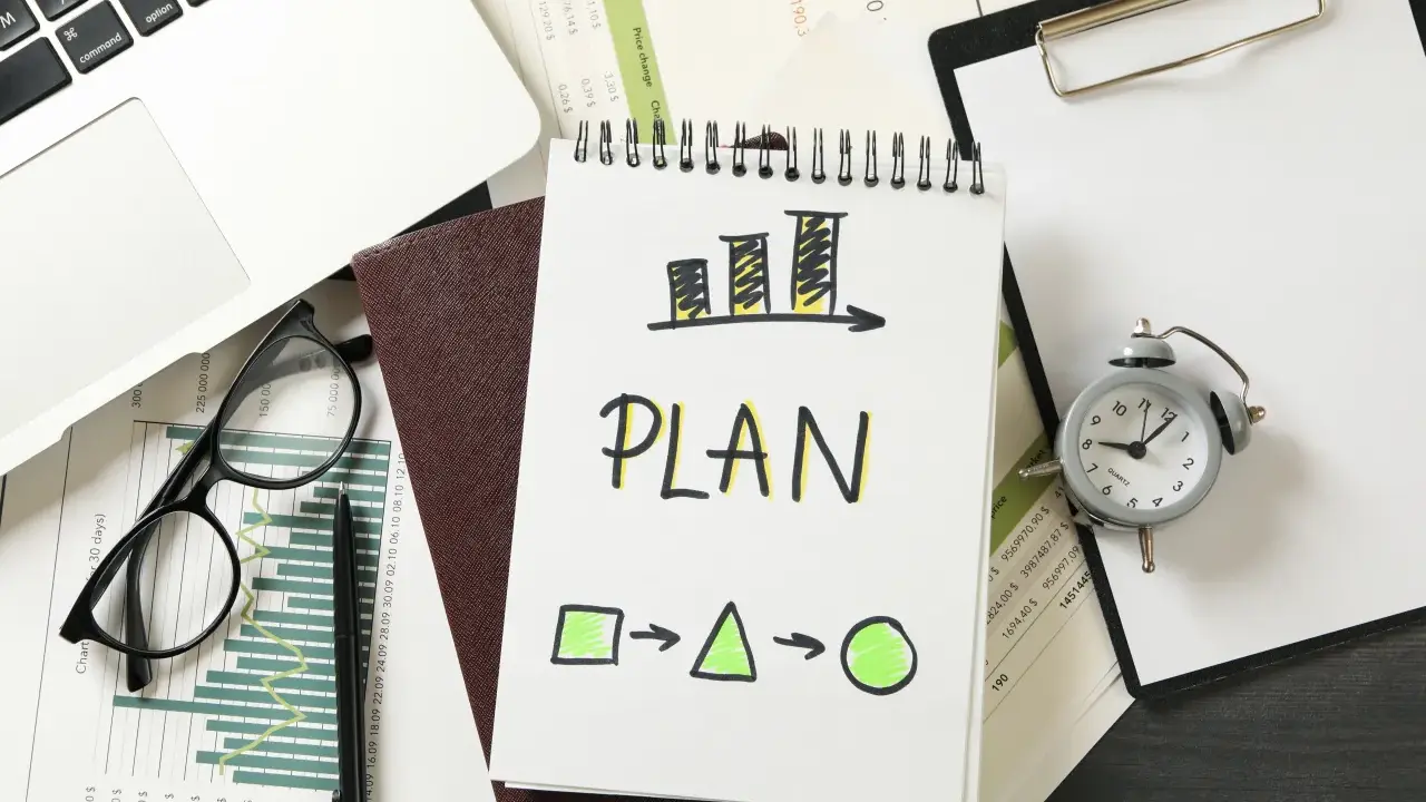 Na biurku leżą dokumenty, laptop, okulary i budzik. Na notatniku widnieje napis "PLAN" i schemat, który symbolizuje indywidualny plan rozwoju.